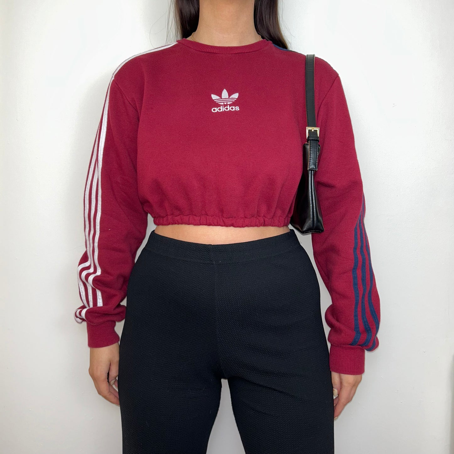 burgundy cropped sweatshirt with white adidas logo shown on a model wearing black trousers and a black shoulder bag