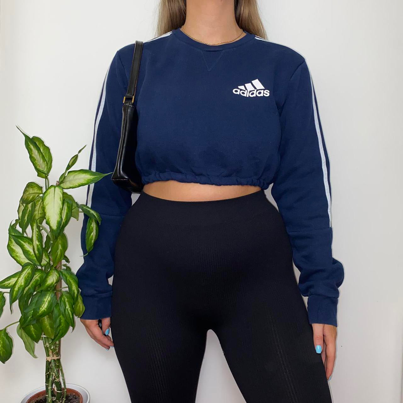 navy sweatshirt with small adidas text and symbol logo on chest with white 3 stripes on arms shown on model wearing black leggings and black shoulder bag