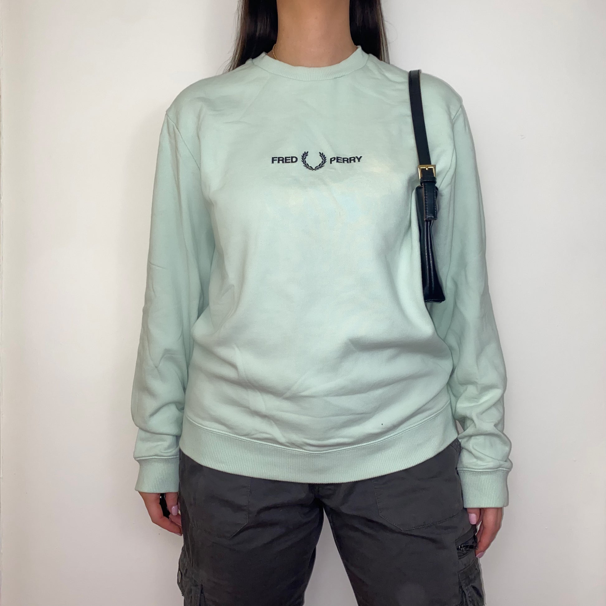 green sweatshirt with black fred perry logo shown on a model wearing grey cargo trousers and a black shoulder bag