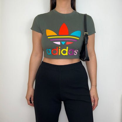 khaki short sleeve crop top with multi coloured adidas logo shown on a model wearing black trousers and a black shoulder bag