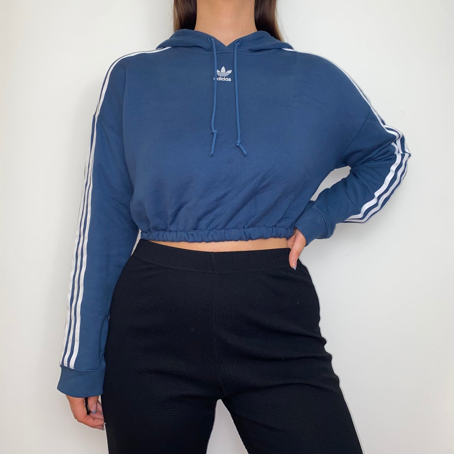 navy cropped hoodie with whtie adidas logo shown on a model wearing black trousers with hand on hip