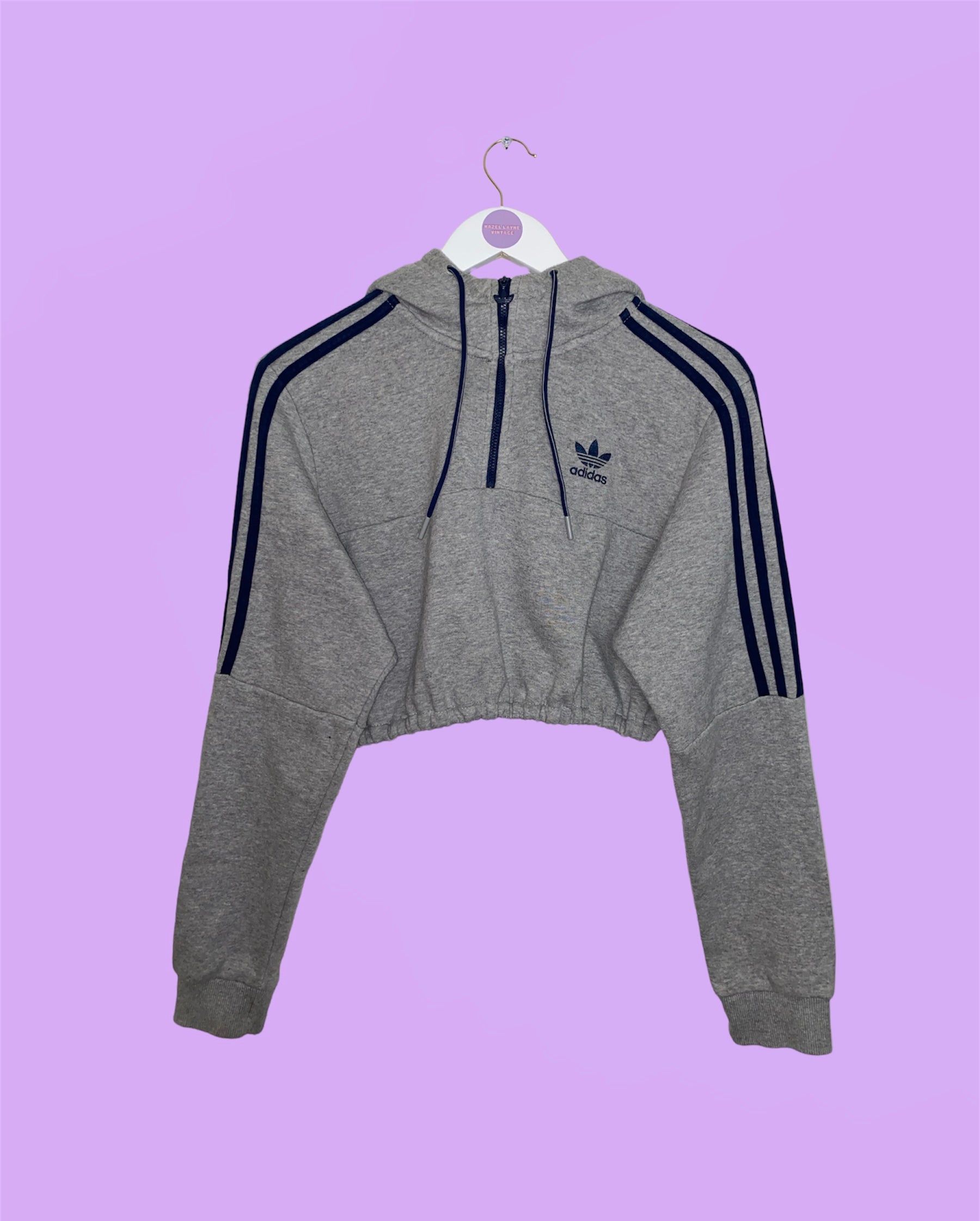 grey 1/4 zip cropped hoodie with navy adidas logo shown on a white clothes hanger on a lilac background