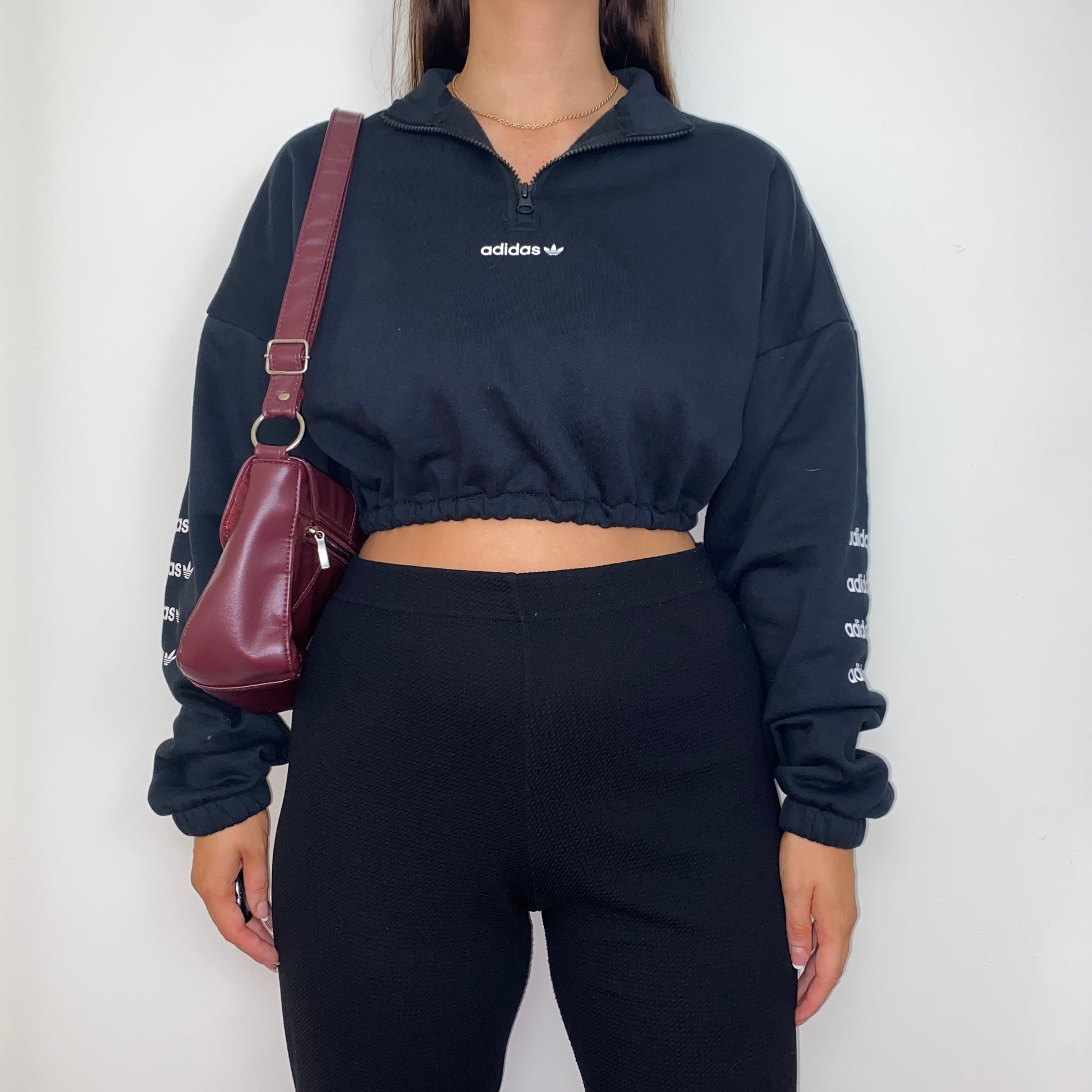 black 1/4 zip cropped sweatshirt with white adidas logo on front and sleeves shown on a model wearing black trousers and a burgundy shoulder bag