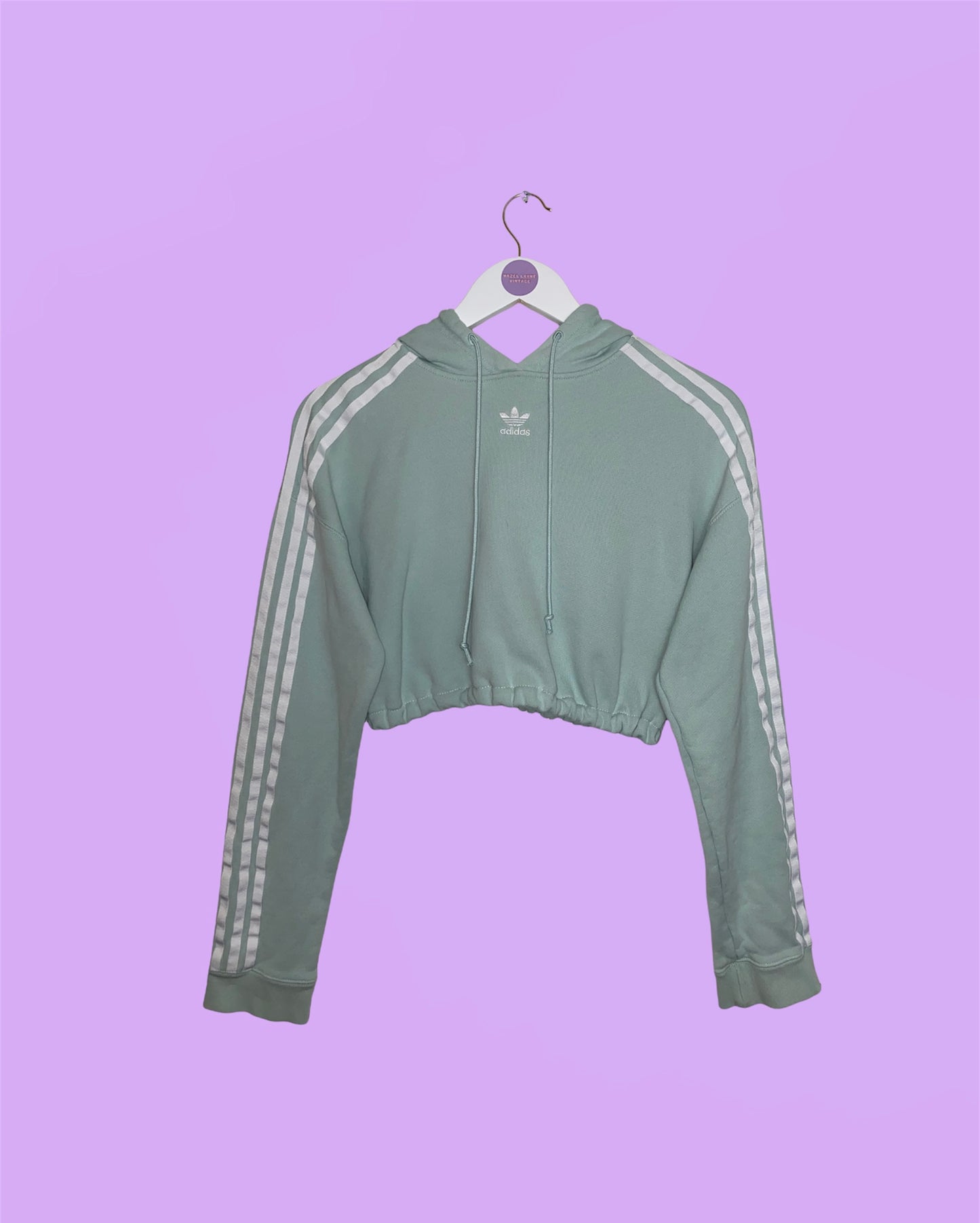 sage green cropped hoodie with white adidas logo and white stripes on sleeves shown on a white clothes hanger on a lilac background