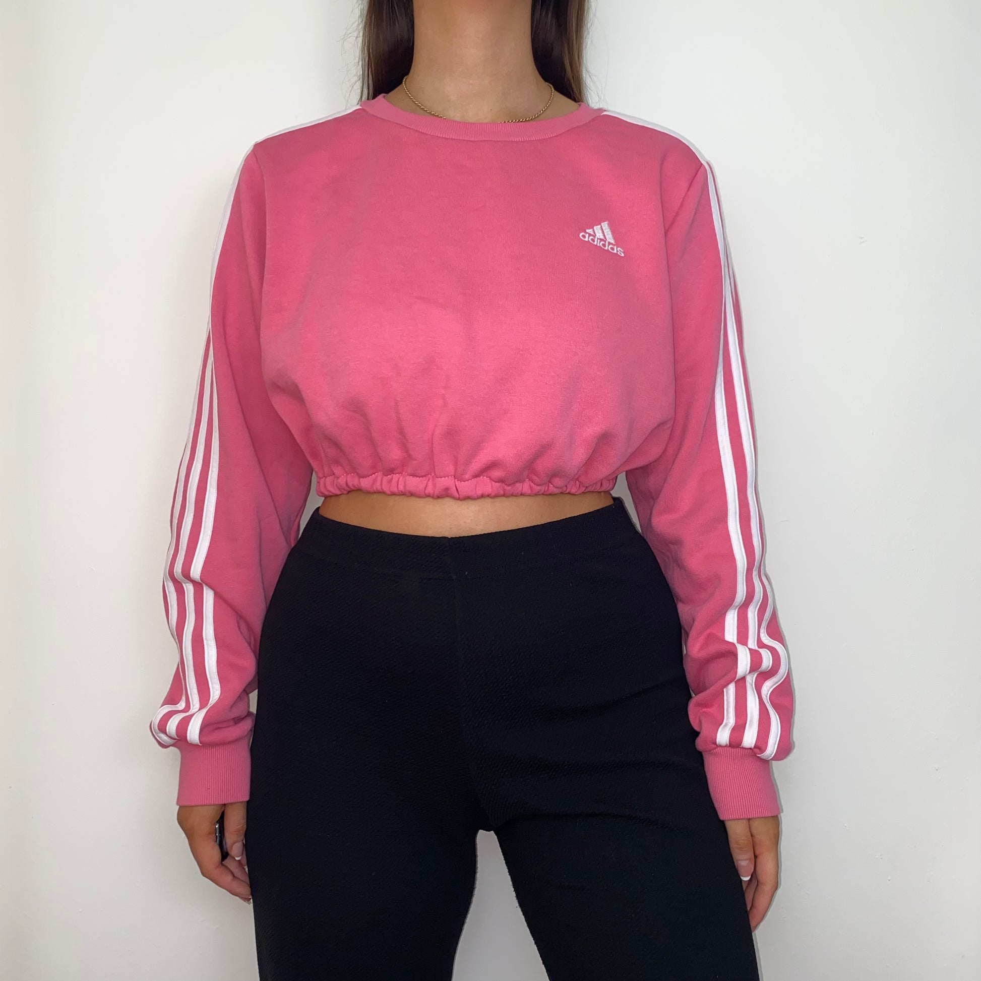 pink adidas cropped sweatshirt with white adidas logo shown on a model wearing black trousers