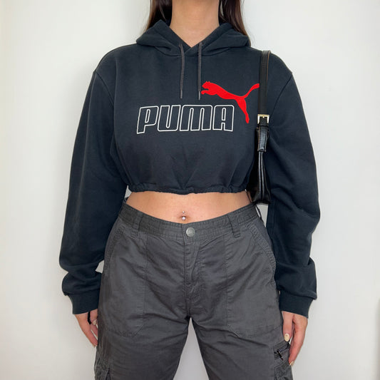 black cropped hoodie with white and red puma logo shown on a model wearing grey cargo trousers and a black shoulder bag