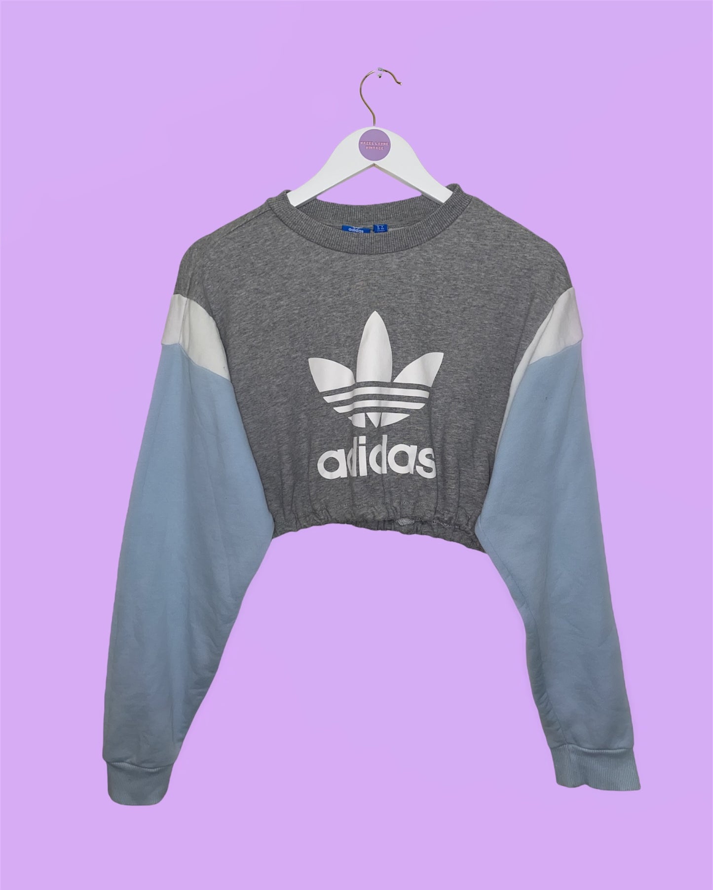 grey cropped sweatshirt with light blue sleeves and white big text and symbol adidas logo shown on a white clothes hanger on a lilac background