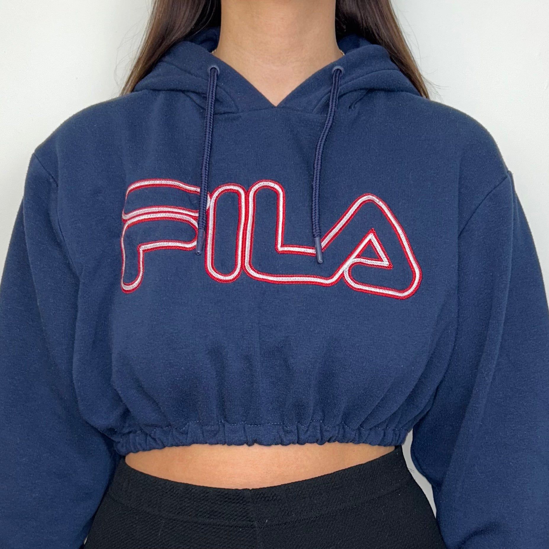 close up of navy cropped hoodie with red big text fila logo shown on a model wearing black trousers