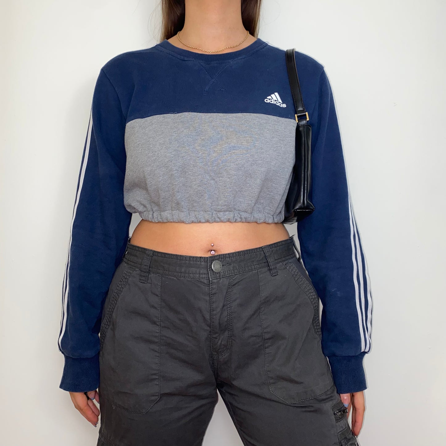 navy and grey cropped sweatshirt with white adidas logo shown on a model wearing grey cargo trousers and a black shoulder bag