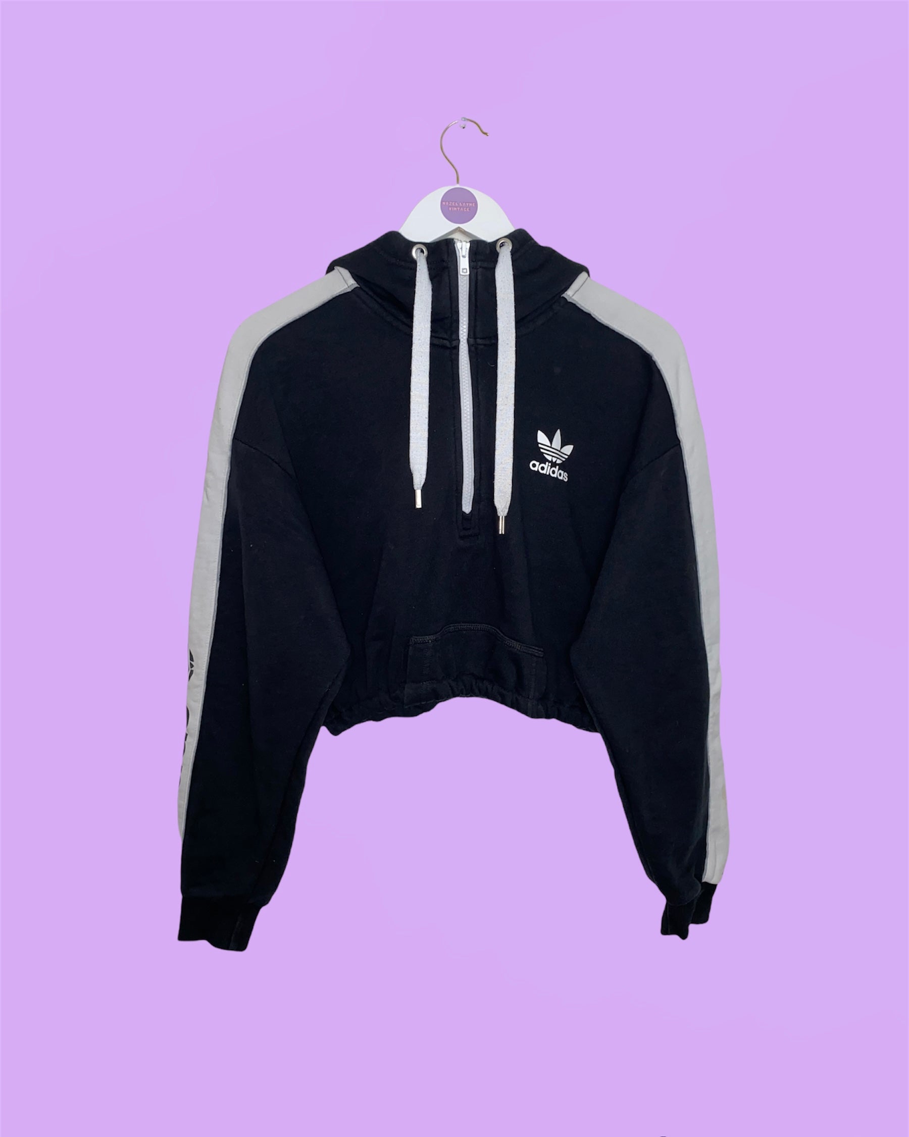 black 1/4 zip cropped hoodie with adidas logo on chest and sleeve shown on a white clothes hanger on a lilac background