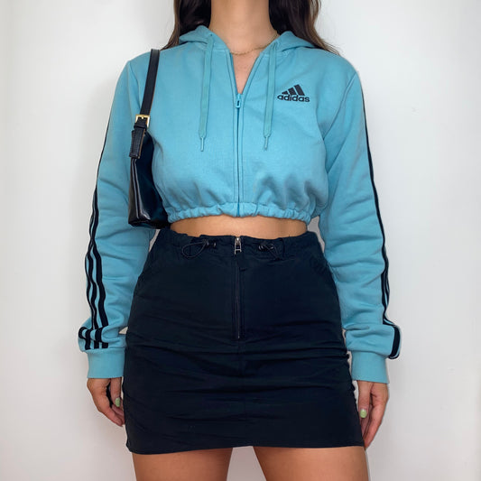 blue zip up crop hoodie with black adidas logo shown on a model wearing a black skirt and black shoulder bag