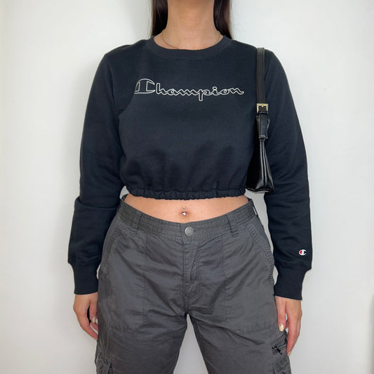 black cropped sweatshirt with white adidas big text logo shown on a model wearing grey cargo trousers and a black shoulder bag