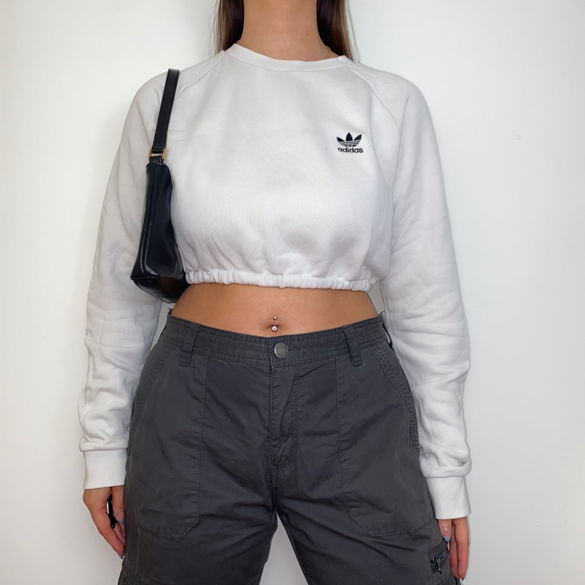 white cropped sweatshirt with black adidas logo shown on a model wearing grey cargo trousers and a black shoulder bag