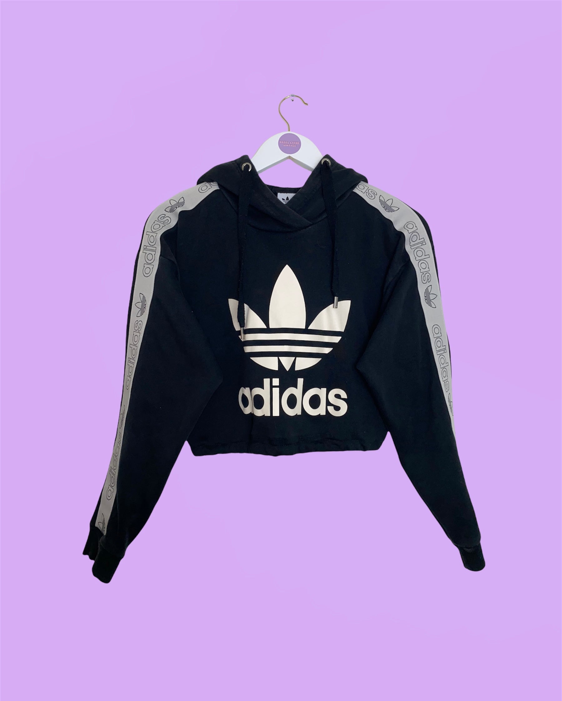 black cropped hoodie with big white adidas logo shown on a white clothes hanger on a lilac background