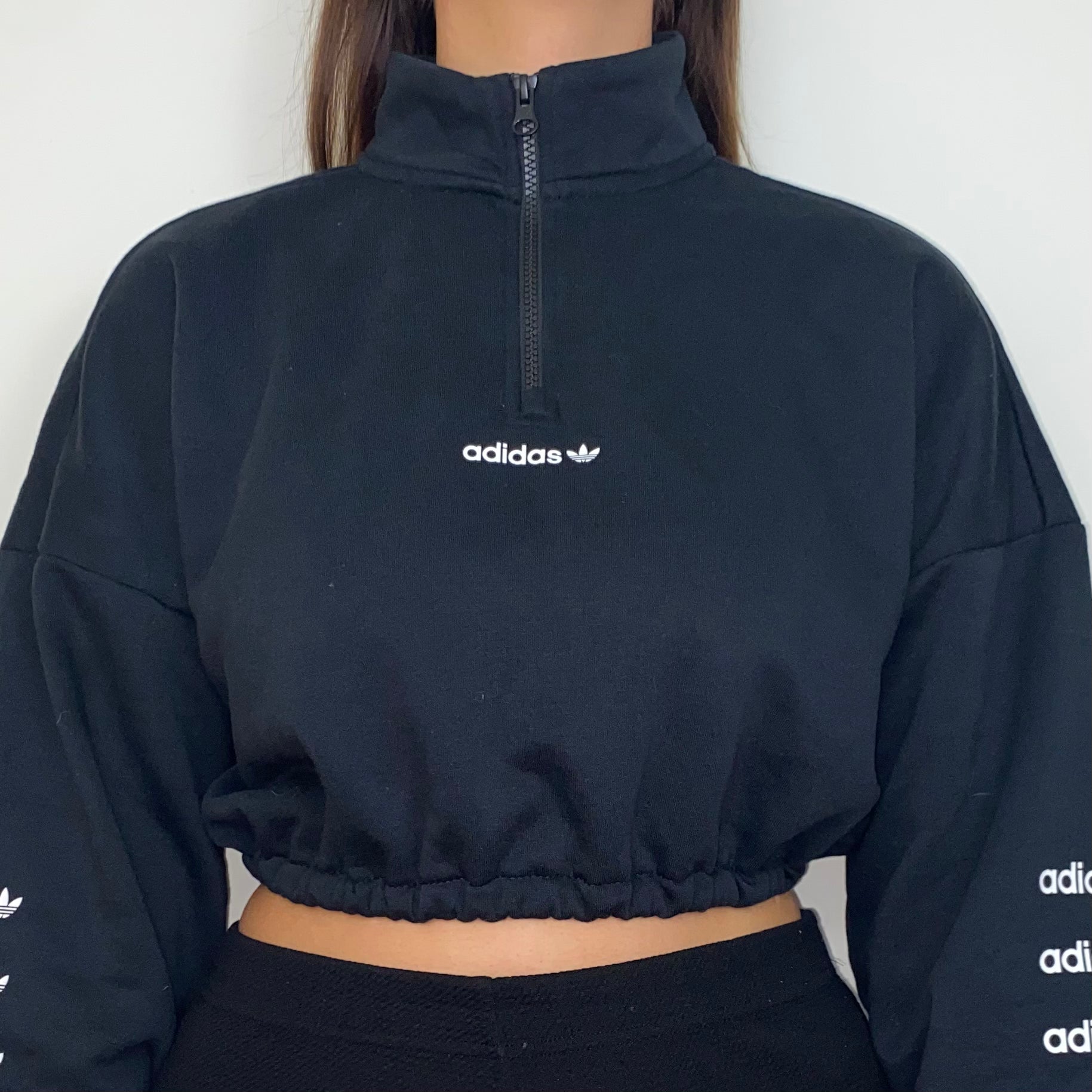 close up of black 1/4 zip cropped sweatshirt with white adidas logo on front and sleeves shown on a model